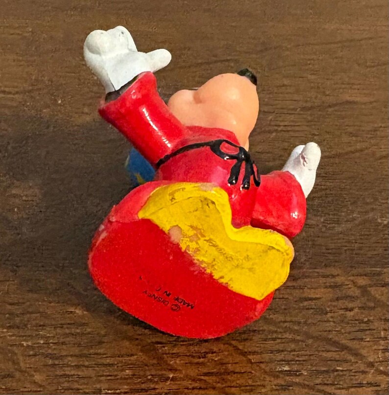 May include: A vintage figurine of a cartoon character in a dynamic pose. The figure is primarily red, with yellow and blue accents. It has white gloves and a black nose. The figurine is on a wooden surface.