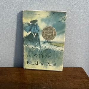 May include: A vintage paperback book titled "The Witch of Blackbird Pond" by Elizabeth George Speare. The cover features a watercolor illustration of a young woman in a blue cloak standing in a field with a pond in the background. The book has a gold John Newbery Medal sticker on the front cover.