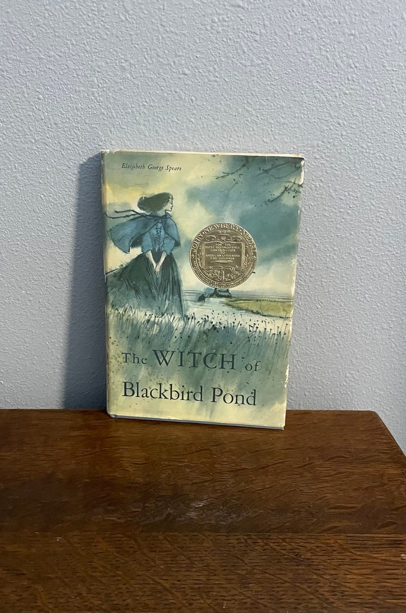 May include: A vintage paperback book titled "The Witch of Blackbird Pond" by Elizabeth George Speare. The cover features a watercolor illustration of a young woman in a blue cloak standing in a field with a pond in the background. The book has a gold John Newbery Medal sticker on the front cover.