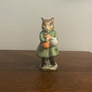 Könnte beinhalten: Keramikfigur einer Katze in einem grünen Mantel, die eine kleine orangefarbene Tasche und einen weißen Behälter hält. Die Katze hat braunes und beiges Fell und steht auf einem Sockel mit strukturiertem Design.