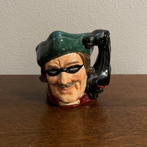 Puede incluir: Una taza de cerámica con forma de cabeza de hombre que lleva un sombrero verde y una máscara negra para los ojos. La taza tiene un asa negra con forma de mano.