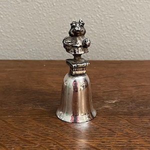 May include: Silver bell with a pig figurine on top. The pig is holding a basket of fruit. The bell is on a wooden surface.