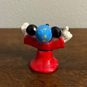 May include: A small figurine of a character in a red robe with black ears and white gloves. The character's head is blue with yellow stars and a crescent moon. The figurine is standing with its arms raised.