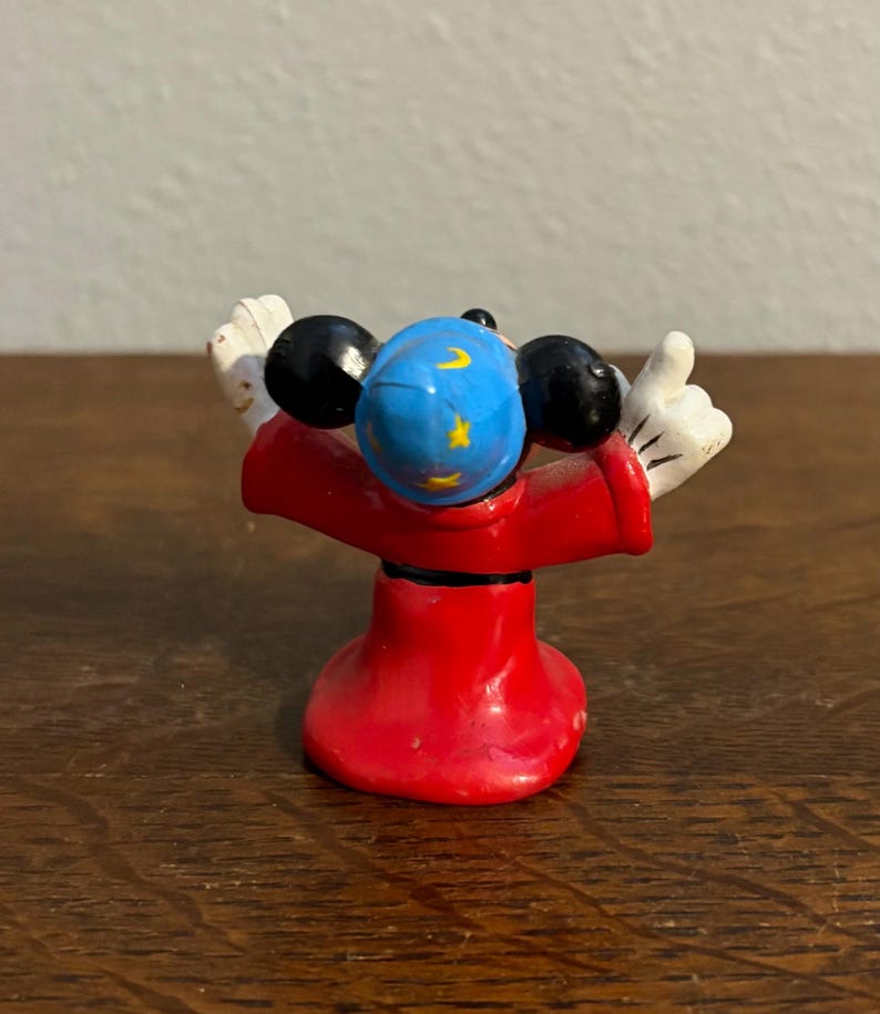 May include: A small figurine of a character in a red robe with black ears and white gloves. The character's head is blue with yellow stars and a crescent moon. The figurine is standing with its arms raised.