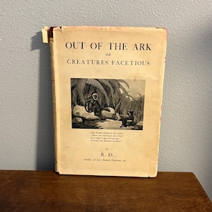 May include: Aged vintage book, "OUT OF THE ARK OR CREATURES FACETIOUS", featuring a black and white illustration. The cover shows signs of wear. The book is displayed on a wooden surface.
