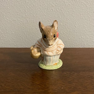 May include: A small ceramic figurine of a brown mouse wearing a striped shirt and holding a basket. The mouse has a pink bow on its ear and is standing on a green base.