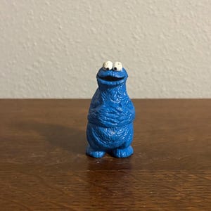 May include: A small, blue Cookie Monster figurine with white eyes and a wide, smiling mouth. The figurine has a textured surface and is standing upright. The toy is a collectible item.