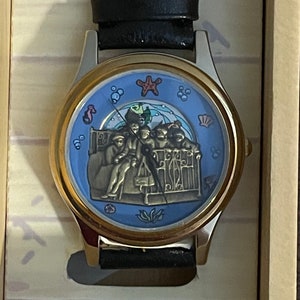 May include: A gold-tone wristwatch with a blue face featuring a nautical scene with a starfish, seashells, and a group of people in a boat. The watch has a black leather band.