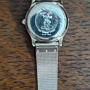 May include: Silver-toned wristwatch with a mesh band, featuring a round face with a Mickey Mouse design. The watch band is made of stainless steel. The watch is displayed on a wooden surface.