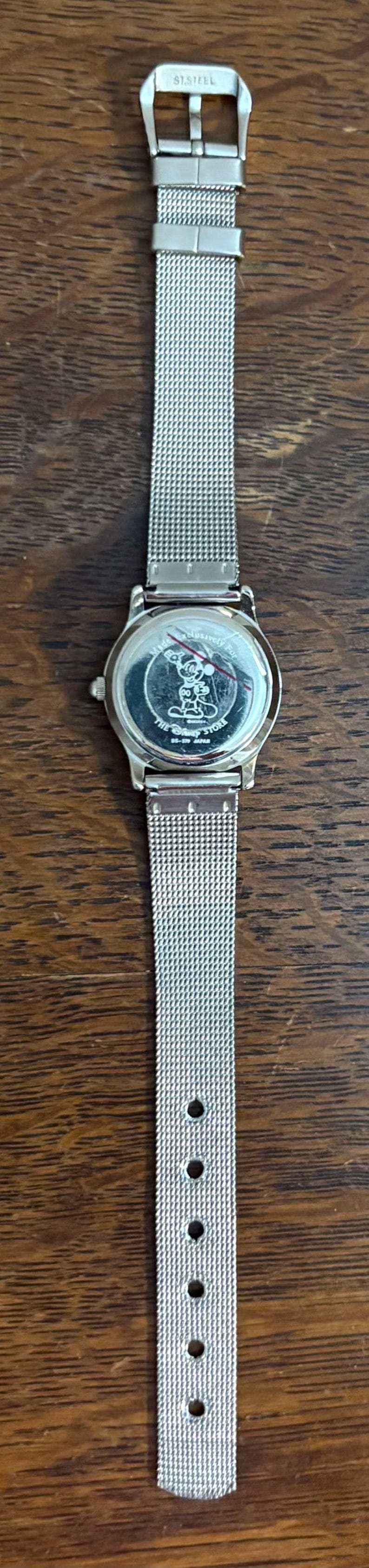 May include: Silver-toned wristwatch with a mesh band, featuring a round face with a Mickey Mouse design. The watch band is made of stainless steel. The watch is displayed on a wooden surface.
