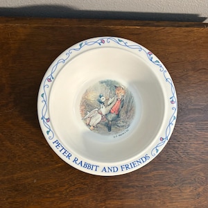 May include: A round white ceramic bowl featuring a central illustration of a duck and a fox. The rim is decorated with a blue vine pattern with small pink flowers. The text "PETER RABBIT AND FRIENDS" is printed in blue along the bottom edge.