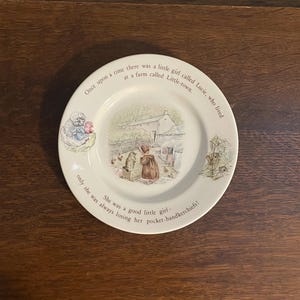 Puede incluir: Un plato de cerámica color crema con una ilustración central de una escena de granja con una niña y un gato. El borde muestra el texto: Once upon a time there was a little girl called Lucie, who lived at a farm called Little-town. She was a good little girl—only she was always losing her pocket-handkerchiefs!