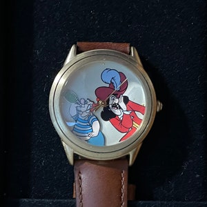 May include: A brown leather wristwatch with a gold-toned case. The watch face features a cartoon image of Captain Hook and Smee from the Disney film Peter Pan.