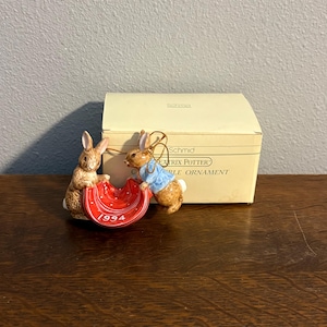 May include: A ceramic Beatrix Potter collectible ornament featuring two brown rabbits holding a red blanket with white polka dots and the year 1994 printed on it. One rabbit wears a blue jacket. The ornament is displayed in front of its original box.
