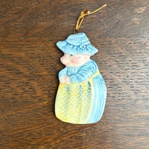 May include: A ceramic pig figurine wearing a blue hat and dress, holding a yellow bag. It has a gold string for hanging. The pig's face is pink, and the dress is light blue and yellow.