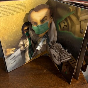 May include: A pop-up book spread featuring a dentist with a green surgical mask and a syringe. The illustration has a vintage style with a radiation symbol and the text "X-30". The book is open on a wooden surface.
