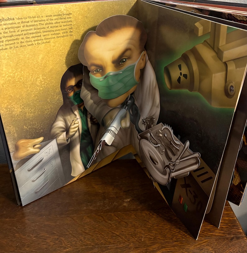 May include: A pop-up book spread featuring a dentist with a green surgical mask and a syringe. The illustration has a vintage style with a radiation symbol and the text "X-30". The book is open on a wooden surface.