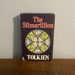 May include: A hardcover copy of "The Silmarillion" by J.R.R. Tolkien. The book has a dark blue cover with the title in red and a circular design in the center. The author's name, Tolkien, is printed at the bottom.