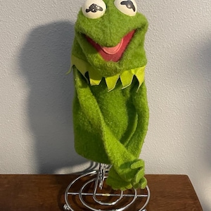 Może przedstawiać: Kermit Żaba kukiełka wykonana z zielonego filcu, z białymi i czarnymi oczami, czerwonymi ustami i zielonym kołnierzem. Kukiełka jest wystawiona na srebrnym metalowym stojaku, na tle jasnoszarej ściany i brązowej drewnianej powierzchni.