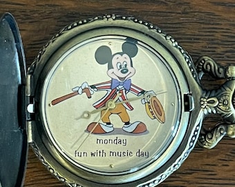 Könnte beinhalten: Eine Vintage-Taschenuhr mit Klappdeckel. Das Zifferblatt zeigt Micky Maus in einem musikalischen Outfit mit den Worten "monday fun with music day". Die Uhr hat ein bronzefarbenes Metallgehäuse und einen dekorativen Griff.