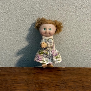 May include: A vintage doll with a smiling face, green eyes, and light brown hair styled in pigtails. The doll wears a floral print dress with purple and white flowers. The doll is standing on a wooden surface.