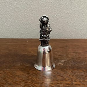 Puede incluir: Campana de plata con una figura de gato en la parte superior. El gato lleva un sombrero y sostiene un objeto pequeño en sus patas.