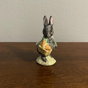 May include: A ceramic figurine of a rabbit holding a basket of carrots. The rabbit is gray with pink-lined ears and wears a green jacket. The basket is yellow and the base is green.