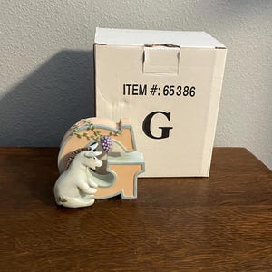 Könnte beinhalten: Dekorative "G"-Buchstabenfigur in Hellpfirsich und Blau, mit einer weißen Eselfigur. Die Figur steht neben einer Box mit dem Aufdruck "ITEM #: 65386". Der Buchstabe ist mit floralen Akzenten und einer Traube verziert.