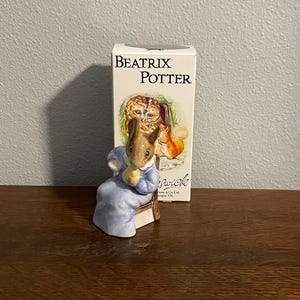 May include: A ceramic figurine of a rabbit in a blue dress seated on a chair, with a box behind it. The box is labelled "Beatrix Potter" and features an illustration of an owl and a squirrel. A charming collectible.