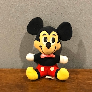 May include: A vintage Mickey Mouse plush toy. The plush has black ears, a yellow face, and a black body with red shorts and white polka dots. It has a pink bow tie and yellow feet. The toy is sitting on a wooden surface.
