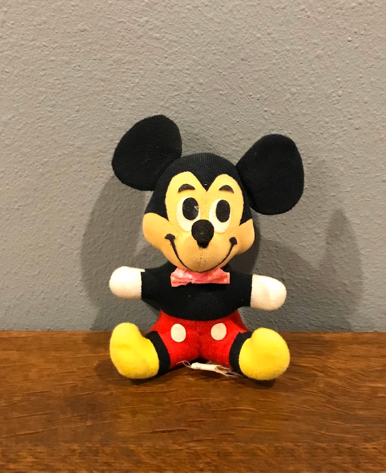 May include: A vintage Mickey Mouse plush toy. The plush has black ears, a yellow face, and a black body with red shorts and white polka dots. It has a pink bow tie and yellow feet. The toy is sitting on a wooden surface.