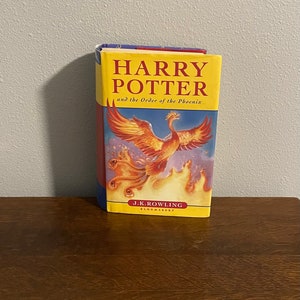 May include: A yellow, red, and blue paperback book with the title "Harry Potter and the Order of the Phoenix" and the author "J.K. Rowling". The cover features a phoenix in flight with flames.