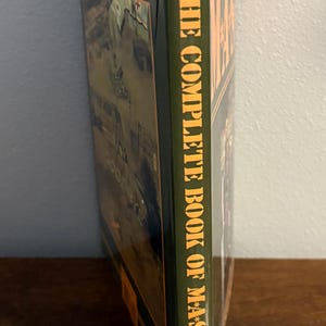 Op de afbeelding: Een hardcover boek getiteld "The Complete Book of M*A*S*H" van Kalter. Het boek heeft een groene kaft met witte tekst en een afbeelding van een militaire sc&egrave;ne. Het boek ligt op een houten oppervlak.