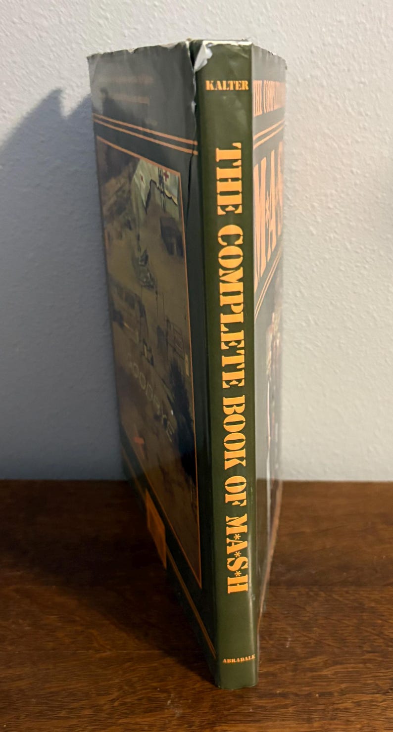 Op de afbeelding: Een hardcover boek getiteld "The Complete Book of M*A*S*H" van Kalter. Het boek heeft een groene kaft met witte tekst en een afbeelding van een militaire sc&egrave;ne. Het boek ligt op een houten oppervlak.