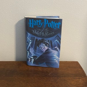 May include: A blue and black paperback book with the title "Harry Potter and the Order of the Phoenix" by J.K. Rowling. The cover features an illustration of Harry Potter in a dark room with a wand in his hand.