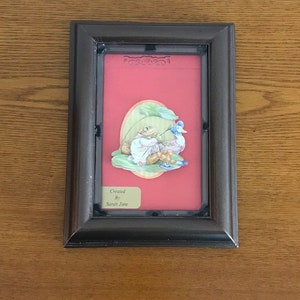 May include: A framed 3D paper craft of a frog and a duck sitting on a lily pad. The frame is brown with a red mat and a gold plaque that reads "Created By Sarah Jane".