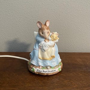 Peut inclure: Veilleuse en céramique représentant une souris mère tenant un bébé souris et un ours en peluche. La souris porte une robe bleue. La base de la veilleuse indique "Baby's First Christmas".