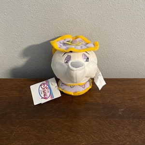 May include: A white plush toy teapot with a yellow lid and purple accents. The teapot has a face with closed eyes and a small spout. The toy is labelled "Disney Store".