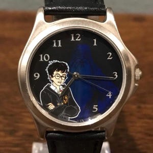 May include: A silver wristwatch with a black watch face featuring a cartoon image of a bespectacled character in a black robe. The watch has a black leather band and silver hands. The watch face also has the numbers 1 through 12.