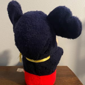 May include: A plush Mickey Mouse toy, viewed from the back, features a navy blue head and ears, a yellow collar, and red shorts. The toy is positioned on a wooden surface against a neutral wall.