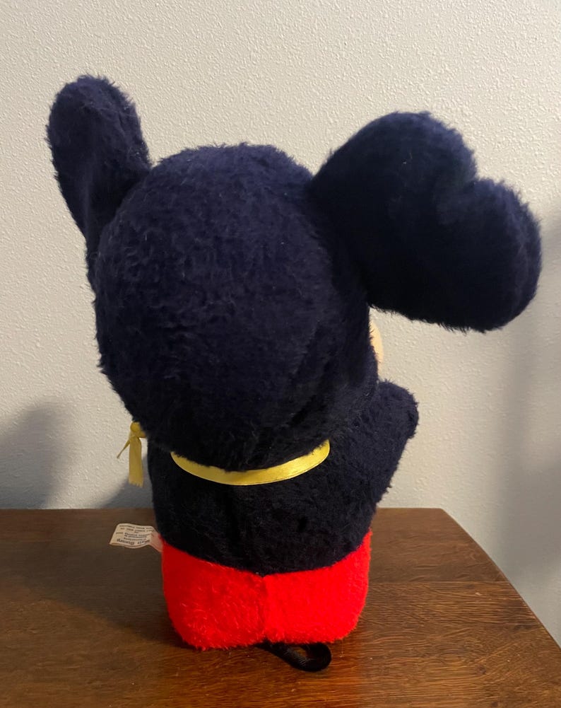 May include: A plush Mickey Mouse toy, viewed from the back, features a navy blue head and ears, a yellow collar, and red shorts. The toy is positioned on a wooden surface against a neutral wall.