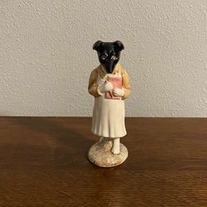 May include: A ceramic figurine of a dog wearing a dress and holding a book that says "CADILLAC".
