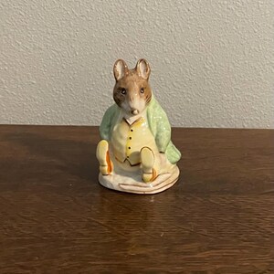 May include: A small ceramic figurine of a mouse wearing a green jacket and yellow shoes. The mouse is sitting on a base and has a brown and white coat.