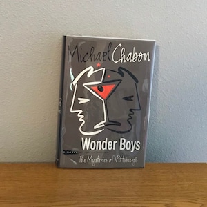 May include: A hardcover book titled "Wonder Boys" by Michael Chabon. The cover is dark gray with a stylized illustration of two faces and a cocktail glass. The book is on a wooden surface.