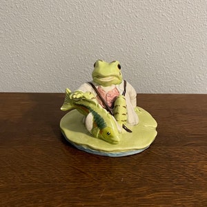 Op de afbeelding: Een groene kikkerfiguur met een wit shirt en een roze strikje zit op een groene waterlelie en houdt een groen en bruin visje vast.