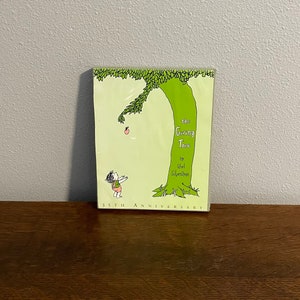 May include: A green book cover with a drawing of a tree and a child reaching for an apple. The title of the book is "The Giving Tree" by Shel Silverstein. The book cover also says "35th Anniversary".
