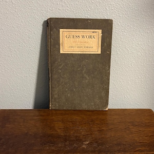 Pode incluir: Um livro antigo intitulado "Guess Work" de Emily Shaw Forman. O livro tem uma capa verde escura com um rótulo retangular com o título e o nome do autor. O livro está sobre uma superfície de madeira.