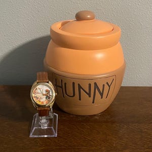 May include: A gold-toned wristwatch with a brown leather band and a colorful face featuring a cartoon character. Beside it is a large, orange and brown ceramic honey pot with the word "HUNNY" in black letters.