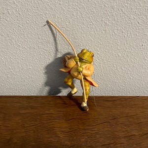 Op de afbeelding: Een groene kikkerfiguur die een geel en groen geruit pak draagt en een hengel vasthoudt. De kikker staat op een houten oppervlak.