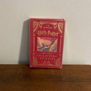 Può includere: Un libro rilegato in rosso con lettere dorate. Il titolo è "Harry Potter e la Camera dei Segreti". Il libro è un'edizione da collezione di J.K. Rowling.
